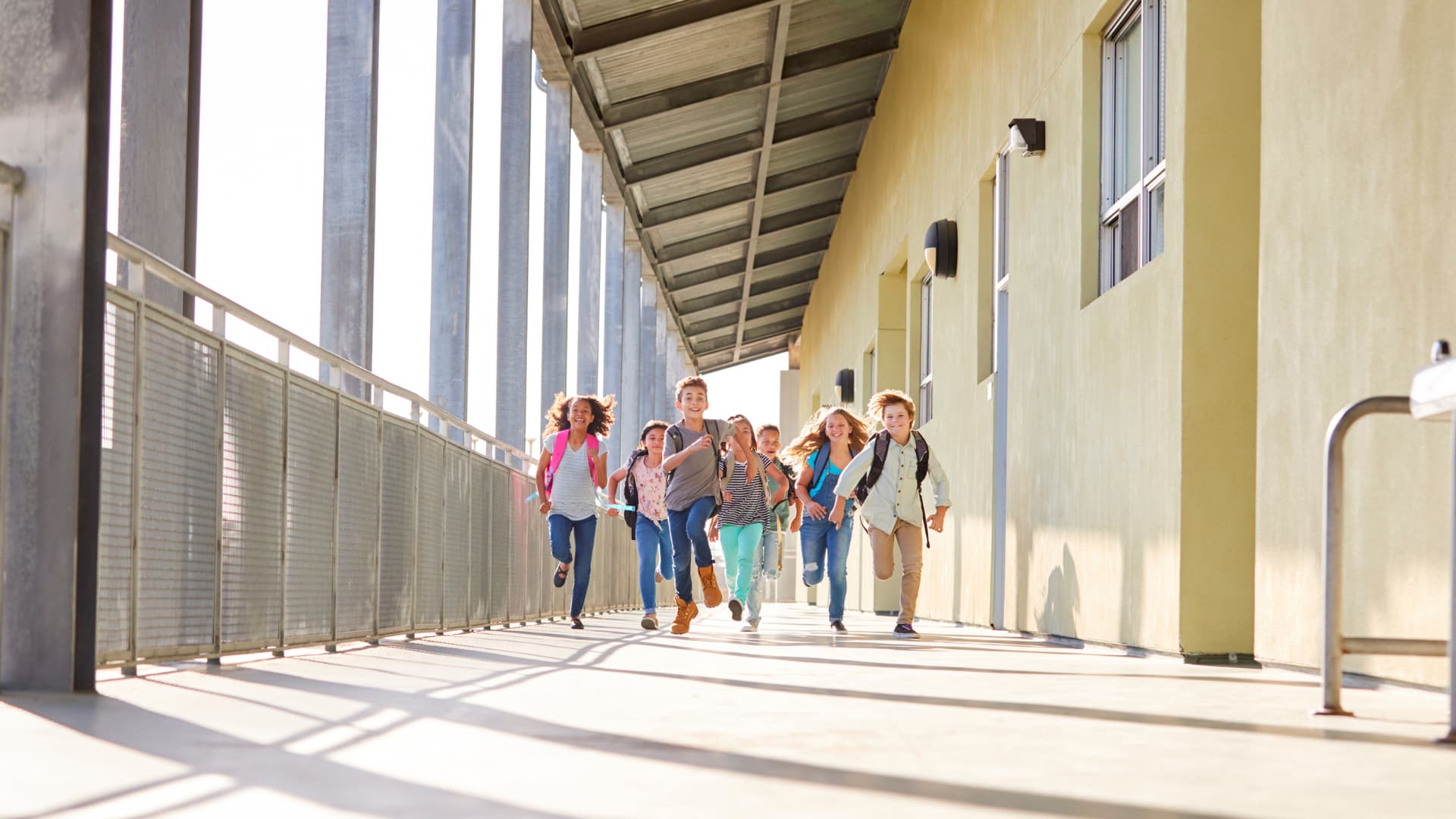 A group of seven children running excitedly down a bright hallway with large windows. They are smiling and wearing casual clothes, carrying backpacks. The sunlight shines through the windows, casting long shadows on the floor.